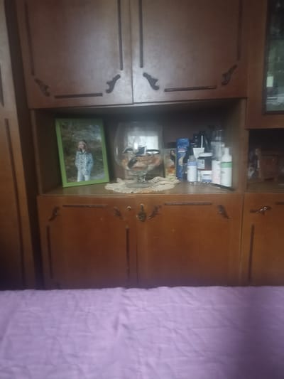 An indoor scene depicts a wooden cabinet with two visible doors on the lower half and two visible doors on the upper half. The upper cabinets have decorative carvings. The lower cabinet doors have ornate handles. The open shelf above the lower cabinets displays a framed photograph of a young girl, a glass bowl with decorative items, and several bottles of toiletries. To the right of the shelf, a framed picture is visible within a cabinet. The scene is viewed from a lower vantage point, with a creased lavender sheet or blanket occupying the bottom portion of the image. The location is identified as Perelešinskij, Russia.