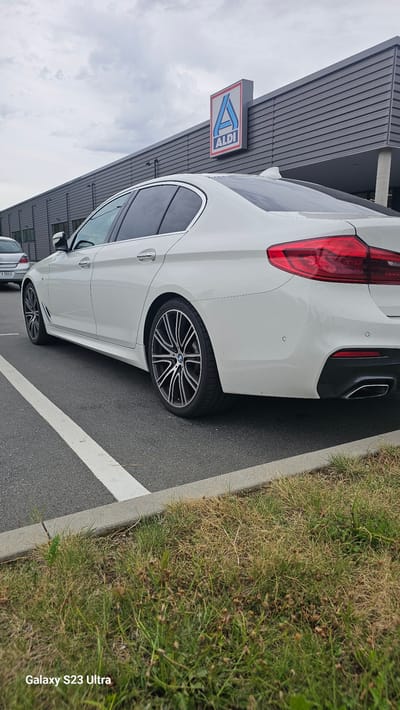 A white BMW sedan is parked in an outdoor parking lot in front of an Aldi store. The car is viewed from the rear passenger side, with the back half of the vehicle and its alloy wheels prominently displayed. The Aldi logo, a red and blue triangular sign with the word "ALDI" underneath, is visible on the building. The parking lot surface is asphalt, marked with white lines. Adjacent to the parking lot is a grassy area with dry, sparse vegetation. The sky is overcast. The license plate of a car in the background is partially visible and appears to read "ZA 1661." The image is watermarked in the lower left corner with "Galaxy S23 Ultra."