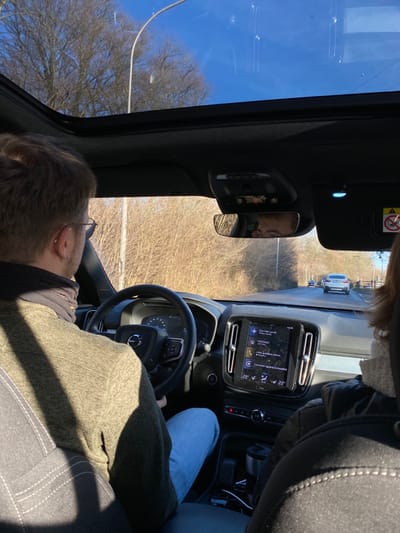 An interior view from a Volvo car, looking forward and slightly to the right, captures the driver, passenger, dashboard, and the external environment under bright daylight in Ans, Belgium.
In the foreground to the left, the back of a person's head, likely the driver, is visible, wearing glasses, with dark hair, and clad in a green sweater over a dark shirt. Their hands rest on the steering wheel, which features a Volvo logo. To the right, a passenger's legs, dressed in blue jeans, are visible. A person's face, likely the passenger with light hair, is reflected in the rearview mirror, looking forward.
The car's dashboard prominently features a vertical infotainment screen. The screen displays the "VOLVO" logo, "Actuellement en cours" (Currently playing), "NOSTALGIA" (likely a music channel or playlist), "Connecter pour compatible" (Connect for compatible), and two temperature readings of "22°". Above the rearview mirror, a sun visor shows an airbag warning symbol. Through the car's large window on the left, a clear blue sky is visible.
Outside the car, the ground is covered with dry, straw-colored vegetation, and numerous bare deciduous trees stand against the bright sky, indicating late winter or early spring. A road or track with a white line is visible in the middle distance, and several other vehicles, including a white van, are parked further away. The car appears to be stationary, possibly parked on a roadside or in a parking area. The visible French text on the infotainment screen aligns with the Belgian location.