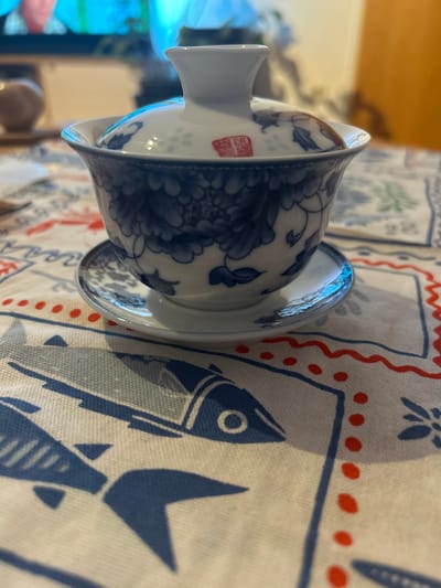 A close-up, eye-level shot shows a blue and white porcelain tea cup and saucer with a lid, resting on a tablecloth. The cup and lid are decorated with intricate blue floral patterns and a small red square stamp with Chinese characters on the lid. The saucer has a similar blue pattern. The tablecloth features a repeating pattern of blue fish and red dots. The background is blurred, showing a television screen with bright colors and a wooden surface on the right. The scene appears to be indoors, possibly in a domestic setting in Hallein, Austria.