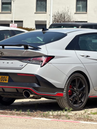 A white car, likely a Hyundai Elantra N due to the visible "N" badge and partial "ANTRA" lettering, is parked outdoors on a paved surface. The car is viewed from its rear-three-quarter angle, showcasing its sporty design with black multi-spoke wheels, red brake calipers, and a distinctive red accent trim running along the bottom of the rear bumper. A single large, circular exhaust pipe protrudes from the right side of the rear. The car has dark tinted windows and a shark fin antenna on the roof.

Next to the parked car, there's a curb with sparse, green vegetation growing along its edge. In the background, a light-colored building with multiple rectangular windows is visible, suggesting a residential or commercial area. Another white vehicle, possibly a van or SUV, is partially visible parked in front of the main car.

The scene takes place during bright daylight, indicating clear and sunny weather. A yellow Israeli license plate, partially visible as "004", confirms the location in Israel, specifically Kfar Monash as provided. A red and white sign in the background displays Hebrew text, "לרכב" (for a vehicle/to ride) and "כביש אש" (fire road/emergency road), suggesting a restricted or emergency access zone. No people are visible in the image.
