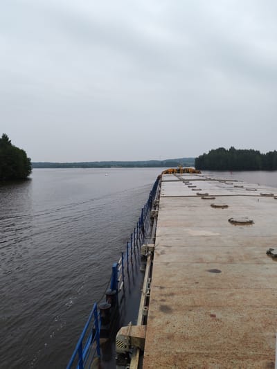 The image depicts a wide river with a barge moving along its surface. The barge's deck occupies the right side of the frame, showing a large, flat, metal surface with numerous circular hatches. A blue railing runs along the edge of the deck. The water on the left side of the frame displays a wake from the barge, indicating movement. Lush green trees line the distant banks of the river, and the sky is overcast and a pale gray. The scene appears to be in a natural, possibly rural, setting. The specific location in Goritsy, Russia is not discernible from the visual information provided. There are no people visible in the image.