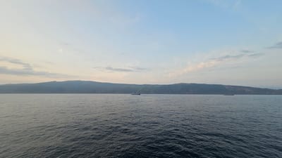 This is a wide-angle shot of a calm sea under a hazy, pale blue sky with wispy clouds tinged with pink and orange, suggesting either dawn or dusk. In the middle distance, a long, dark cargo ship sails horizontally across the frame. Further in the distance, a hilly, green coastline forms a long, undulating horizon line, with some visible structures or radio towers on its crest. Another ship, much smaller and farther away, can be seen near the right edge of the frame. The water is dark blue and shows gentle ripples. The overall mood is peaceful and serene. The image captures a moment of quiet transit in a coastal setting. The lack of clear shadows and the soft light suggest a time of day away from direct sunlight. The visible features of the landscape, particularly the mountainous coastline, are consistent with a location like Pasaia, Spain, known for its dramatic coastal geography and maritime activity. There is no visible text.