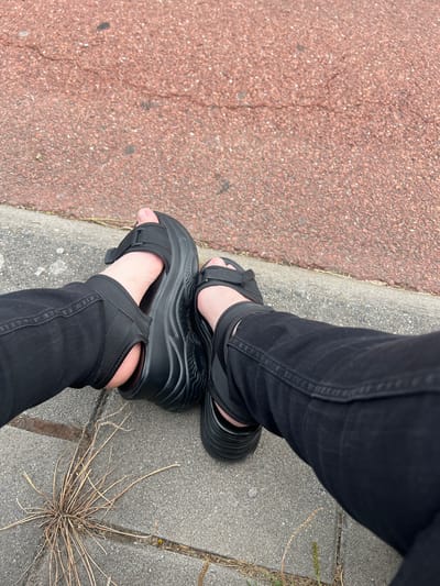 This media file shows a close-up of a person's legs and feet, adorned with black platform sandals and black jeans. The individual is standing on a paved surface, with a reddish-brown, textured area to the left, likely a sidewalk or path. The background consists of gray concrete slabs, with a few dried sprigs of grass growing in the cracks. The image is taken from a top-down perspective, focusing on the footwear and lower legs. The lighting suggests it is daytime, and the overcast appearance of the ground might indicate a cloudy day. There are no visible text elements or indications of a specific event or strong emotions. The setting appears to be an urban or suburban outdoor environment, possibly a street or park. The Netherlands is provided as a location context, suggesting this scene could be in Leeuwarden.