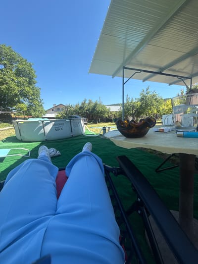 In Suvorovo, Bulgaria, an individual is reclining on a chair outdoors, enjoying a sunny day under a covered patio. The scene unfolds in a backyard setting, likely a residential area, given the presence of houses and trees in the background. The ground is covered with artificial turf, and a Bestway Steel Pro MAX above-ground pool is visible.

The person is wearing light blue trousers and white sneakers. Their perspective is from a low angle, looking upwards and outwards. On a table nearby, there's a glass of water with ice and a slice of lemon, a pack of cigarettes, and an amber-colored decorative bowl. The table is situated beneath a white, corrugated metal roof supported by dark metal poles, providing shade. Trees, some with green foliage and others with yellowing leaves, dot the landscape, suggesting it might be late summer or early autumn. The sky is clear and bright blue. The overall impression is one of relaxation and leisure in a suburban or rural outdoor space.
