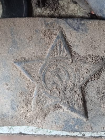 A close-up, eye-level, outdoor shot shows a dusty, embossed five-pointed star with a hammer and sickle emblem inside, on a dark, textured surface. The star and emblem are worn and partially obscured by a layer of light-colored dust and debris. The background is out of focus, showing a dark, possibly metallic object at the top and a lighter, textured surface at the bottom. The scene appears to be located on the ground in Žygaičiai, Lithuania, given the metadata. There are no people or actions depicted in the image. The lighting is even, suggesting it is daytime.