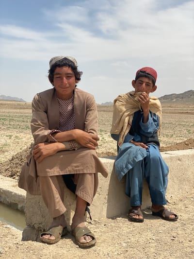 Two young men are sitting on a concrete structure in a rural area. The man on the left wears a brown outfit and a white cap. The man on the right is in a blue outfit and a red cap, with a light-colored shawl draped over his shoulders. He appears to be eating something he is holding in his hand. The background features a dry landscape and distant hills under a blue sky. Based on the landscape and the clothing of the men, this image likely portrays a scene in or around Ashoqah, Afghanistan.