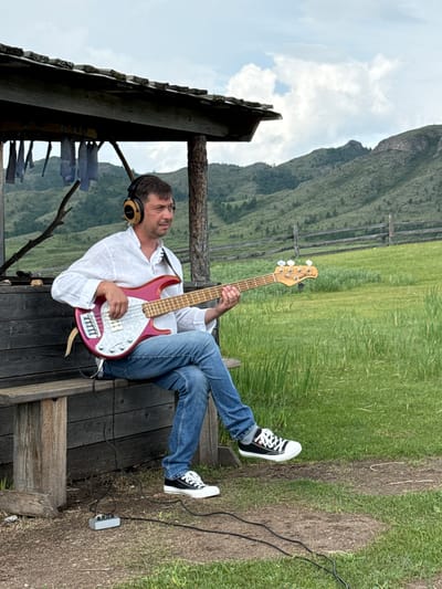 A man with short brown hair and a white collared shirt is sitting on a wooden bench, playing a red electric bass guitar. He is wearing blue jeans and black and white sneakers. He has headphones on and is looking to the right. The bench is part of a rustic wooden structure with a thatched roof, likely an outdoor shelter. The background features a grassy field with a wooden fence and rolling, tree-covered hills under a cloudy sky. The scene appears to be in a rural or mountainous area, possibly near Kazanovka, Russia, given the landscape. A small grey electronic device with cables is on the ground near his feet.