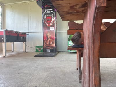 In Glufishevo, Bulgaria, a young boy is seen asleep under a wooden table. His head rests on a dark cushion, with his hair partially visible. In the background, a foosball table stands to the left, and a boxing arcade game featuring Mike Tyson is positioned further back. A green crate is visible near the boxing game. The scene appears to be an outdoor or semi-outdoor recreational area, with a concrete floor and a light-colored wall behind the game.