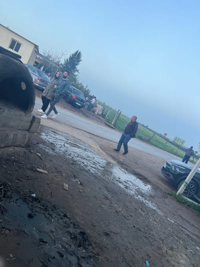 An outdoor scene shows a slightly tilted perspective of a muddy road with several parked cars. A man in dark clothing walks along the road, while a couple stands closer to the viewer. A cow is present in the background. The foreground is dominated by mud and some concrete structures. The sky is overcast, and the overall setting appears to be rural.