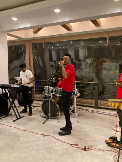 This image captures an indoor musical performance scene in Murera ward, Kenya. Four individuals are present with musical instruments. In the foreground, center-right, a male vocalist with styled dark hair, wearing a red long-sleeved shirt, black pants, and black boots, holds a black microphone to his mouth with his right hand and a small black device in his left. To his left, a male keyboardist, wearing a white t-shirt, black pants, black shoes, and a dark fedora hat, plays a black electronic keyboard positioned on a stand. Behind the vocalist, a drummer, wearing a red shirt, is partially visible operating a black drum kit. On the far right, a partially visible individual in a red shirt and light-colored shoes stands next to a wooden xylophone-like instrument with yellow bars, holding a mobile phone in their left hand. The floor is constructed of light-colored square tiles, across which red and black audio cables are routed. The background consists of a large glass partition, partially covered by transparent plastic sheeting, showing reflections of interior lighting and indistinct forms beyond. The ceiling is light-toned with visible structural beams and integrated recessed lighting fixtures.