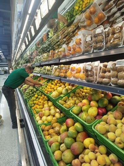 The image shows the produce section of a supermarket in Barquisimeto, Venezuela. A man wearing a green shirt and dark jeans is standing at a display of fruits. He is reaching toward the fruit, appearing to be selecting items. Fruits are arranged in green plastic crates: mangoes, peaches, passion fruits, and oranges are all visible. Above the crates are shelves displaying packaged fruits such as kiwis and pears. Fluorescent lights illuminate the aisle. Price tags are visible on the shelves. The supermarket appears to be well-stocked and brightly lit. Other shoppers can be seen in the background.