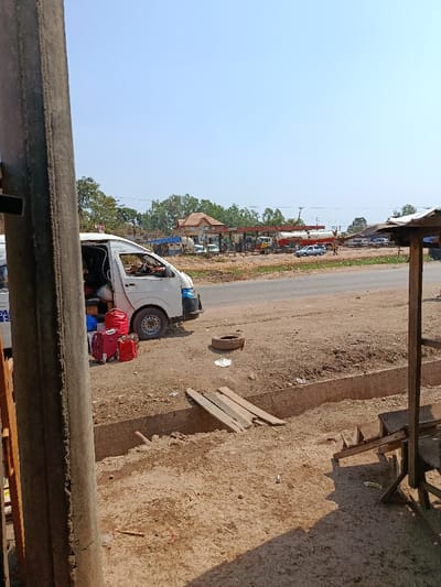 An outdoor scene in Ozalla, Nigeria, depicts a roadside environment on a clear, sunny day. In the foreground, a large, dark vertical post frames the left side of the image. To its right, a white passenger van, possibly a Toyota Hiace, is parked on a dry, light brown dirt shoulder. The van's side sliding door is open, revealing visible items within the interior, and three red pieces of luggage are placed on the ground next to the open door. A tire rim lies flat on the dirt near the van. In front of the van, a concrete drainage ditch runs parallel to the road, with several wooden planks laid across a section, forming a makeshift bridge.
A paved asphalt road traverses the midground. Across the road, a gas station complex is visible. It features multiple red fuel pumps beneath canopies, various vehicles including at least two tanker trucks (one white, one red labeled "PEES"), and a two-story building with a light brown tiled roof. Several indistinct human figures are present near the pumps, suggesting activity. To the far right, a weathered wooden structure with a corrugated metal roof stands on the dirt. The background consists of a clear, light blue sky and scattered green trees.