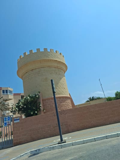 This media file captures a close-up, low-angle view of a cylindrical, stone tower with a crenellated top. The tower is constructed from pale, rectangular stone blocks. To the right of the tower is a reddish-brown brick wall, which extends upwards and to the right. A dark, horizontal pipe or pole runs across the image, partially obscuring the tower and the brick wall.

Below the tower and behind the brick wall, a modern building with multiple windows is visible, along with a lush green bush with white flowers. The foreground shows a paved walkway with blue lines, bordered by a curb and an asphalt road. Several signs are affixed to a metal fence at the bottom right, including what appears to be a safety notice with icons.

The sky is a clear, bright blue, suggesting a sunny day. The lighting indicates it is daytime, likely morning or afternoon. The scene is set in an urban or semi-urban environment, possibly a public area or a courtyard, given the walkway, road, and signage. The architectural style of the tower hints at a historical or decorative function, while the surrounding modern elements suggest a contemporary setting. No people are visible in the frame. The overall impression is one of a bright, clear day in a location with a mix of historical and modern architecture. Visible text includes signage with icons and possibly some graffiti on the brick wall. The location is identified as San Javier, Spain.