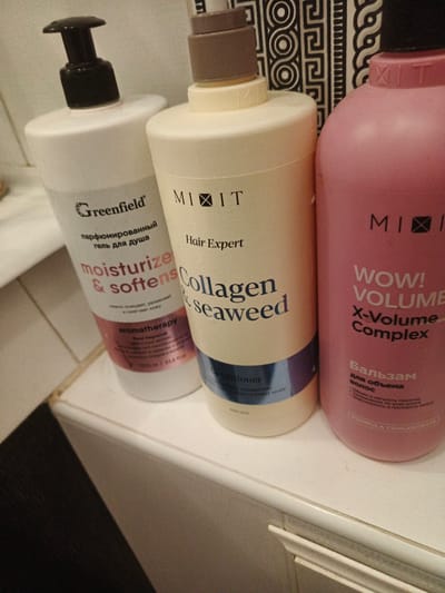 Three bottles of hair care products are arranged on a white surface, likely a countertop or shelf, next to a decorative wall element with a black and white geometric pattern. The bottle on the left is white with a black pump dispenser and features Russian text that translates to "Greenfield, perfumed shower gel, moisturize & softens, aromatherapy." The middle bottle is a light cream color with a brown pump dispenser and displays the brand "MIIT" and English text: "Hair Expert, Collagen & Seaweed, conditioner." The bottle on the right is pink and has a black pump dispenser, with the brand "MIIT" visible. This bottle has Russian text indicating it is a "Balm for volume hair" and English text "WOW! VOLUME X-Volume Complex, FORMULA CONCENTRATE". The lighting suggests an indoor environment, possibly a bathroom or storage area. There are no people visible in the image. The overall scene is clean and organized, showcasing the products. The image does not contain information that would allow for a precise location within Usolye-Sibirskoye, Russia.