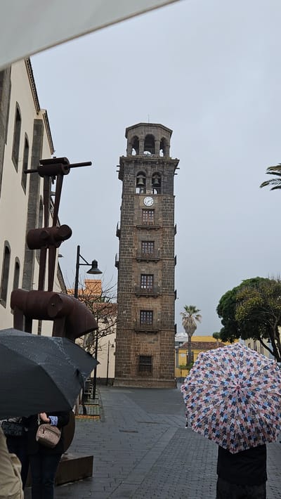 The image depicts an overcast day in San Cristóbal de La Laguna, Spain, focusing on a tall, historic bell tower and people with umbrellas in a paved urban setting.
The main subject is a prominent, dark stone bell tower standing tall on the left side of the frame. It is constructed from large, rectangular blocks and features an upper section with arched openings housing bells. Below the bell section, a white clock face with black Roman numerals is visible, indicating approximately 11:45 or 11:50. Further down the tower are several rectangular windows with dark grates.
In the foreground and mid-ground on the left, an abstract sculpture made of rusty, dark metal is partially visible, featuring industrial-looking components. The ground throughout the visible area is paved with rectangular gray stones, suggesting a pedestrian street or plaza.
On the right side of the image, people are seen walking under umbrellas, indicating overcast or rainy weather. One person prominently holds a colorful umbrella featuring a distinctive pattern of numerous small cats in shades of pink, blue, brown, and white. Another person further back is partially visible under a plain black umbrella.
Behind the pedestrians, other traditional buildings with light-colored walls and dark roofs are present, along with green trees and palm trees, all set against a uniform, light grey sky. The scene captures a moment in a historic town during potentially damp weather. No specific text is clearly readable apart from the Roman numerals on the clock face.