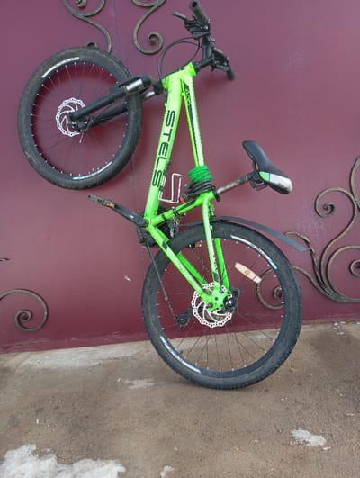 A neon green STELS mountain bicycle is depicted positioned against a dark red vertical surface, identified as being in Yaromaska, Russia. The bicycle features a black seat, black handlebars, and black components including a front suspension fork and disc brakes on both front and rear wheels, identifiable by their silver rotors. A white wire-frame water bottle cage is affixed to the downtube, and a green coiled cable lock is wrapped around the seat tube. A black rear fender is present above the rear tire. The dark red background surface exhibits black wrought iron scrollwork decoration. The ground beneath the bicycle is a rough, light gray concrete or asphalt surface, showing areas of discoloration and dirt. The bicycle's rear wheel rests on this ground, while the front wheel is elevated against the red surface. No individuals are present in the scene.