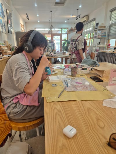 A young female, wearing a gray t-shirt, a pink apron, and black headphones with a white star emblem, sits at a wooden table in an art studio. She is focused on painting a small blue object held in her left hand with a brush in her right hand. The table is covered with a yellow cloth and displays art supplies including paintbrushes, a palette with various colors, small white figurines, and a basket of paints. A person in the background, wearing a striped shirt and an apron, is also visible. The studio environment features artwork on the walls, shelves with art supplies, and large windows allowing natural light. The setting is identified as an art studio in Da Nang, Vietnam.