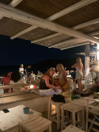The image depicts an outdoor bar or restaurant scene at night in the city of Φάρος, Greece. Artificial lighting illuminates the immediate area, while the background reveals dark hills or landforms against a dark night sky.
The primary structure consists of a thatched roof supported by white-painted wooden beams and crossbeams. Below the roof, the establishment features white-painted, rustic-style wooden furniture. In the immediate foreground, a white wooden table holds a black smartphone and a light blue cylindrical container. Adjacent to this, a white wooden stool is visible. To the right, another white wooden table displays a green glass bottle, a dark object (possibly a small bag or case), and a rectangular wooden box. Two white wooden stools are also present near this table.
In the midground, a low, white-painted bench-like structure with white cushions runs horizontally. Two women are seated on this bench: one with blonde hair, wearing a black tank top and white trousers, is focused downwards, seemingly manipulating an object or food with her right hand. Beside her, another woman with blonde, curly hair, dressed in a light-colored sleeveless top and a yellow skirt, is seated facing her, observing. A small, glowing light source is positioned on the bench near them.
Further to the right, a woman with long blonde hair, wearing a light pink top and light-wash denim shorts, stands with her back partially to the viewer, her right arm raised, suggesting she is drinking from a glass. Near her, a potted plant with spiky green leaves rests on a white stand.
Throughout the midground and background, approximately 15-20 individuals are visible, seated and standing at various white-painted tables and chairs, engaged in social interactions. One individual in a dark shirt is seen standing and moving in the mid-distance. The overall scene suggests a relaxed, social gathering.