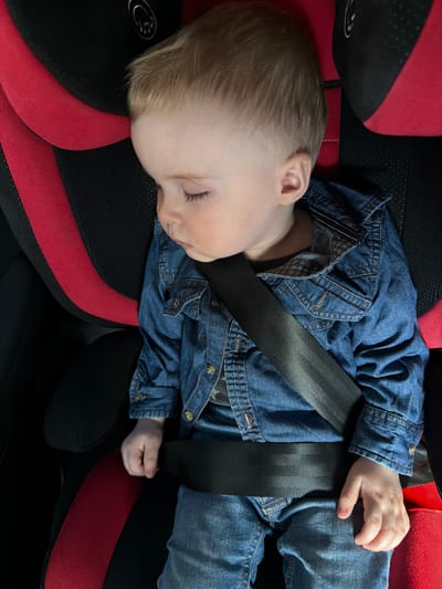 A young child with light blonde hair is observed sleeping in a child safety seat within a vehicle in Bolgury, Russia. The child, wearing a blue denim jacket over a dark shirt and blue jeans, is secured by a black safety harness across the chest and lap. The car seat is predominantly black with red accents on the side bolsters and backrest, featuring a silver-colored emblem on the upper left portion of the headrest. The child's eyes are closed, and the head is tilted forward.