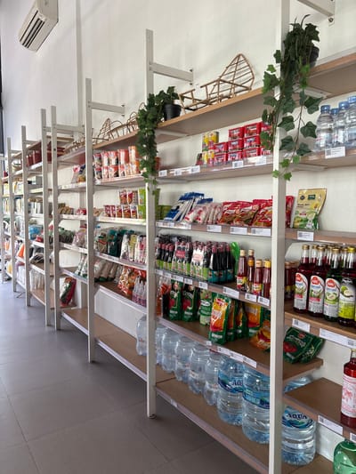 The image depicts the interior of a small grocery store, likely a minimarket, located in Gajahan, Indonesia. The store features multiple shelves stocked with various food items, drinks, and household goods. On the far left, an air conditioning unit is mounted high on the wall. The shelves are made of metal frames with wooden planks.
The shelves on the left are filled with items such as canned goods, packaged snacks, and bottled drinks. Further to the right, the shelves display a variety of packaged foods, including instant noodles, seasonings, sauces, and rice. There are also several bottles of water, including brands like "Le Minerale" and "Aqua," prominently displayed on the lower shelves. Decorative artificial vines are draped from the upper shelves on the right side of the image, adding a touch of greenery to the store's interior.
The overall scene is well-lit and organized, suggesting a typical retail environment. No people are visible in the image. The flooring is tiled with a neutral gray color. The store appears to be a local establishment catering to everyday needs.
