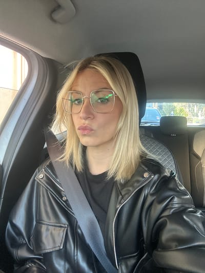 A person with blonde hair, wearing glasses, a black t-shirt, and a black leather jacket, is seated in a car. The person is wearing a seatbelt. Outside the car window, a street scene is visible, possibly located in a city based on the buildings and signage. The location cannot be definitively verified as Vallauris, France, from the image alone.