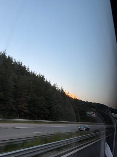 The image depicts a highway scene with a forested hillside on the left and a tunnel entrance on the right. A blue car is driving on the highway. The sky is a pale blue with some hazy clouds. There is a billboard on the hillside. The image appears to be taken from a moving vehicle, as indicated by the motion blur. The specific location in Rajana, Bulgaria, is not identifiable from the image alone.