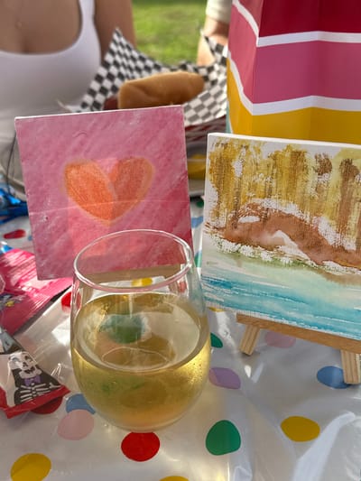 The image captures a close-up, elevated view of a table during an outdoor gathering. The specific location in Accord, United States, is not discernible from the image.
In the foreground, a clear, stemless glass containing a pale yellow liquid and a lime wedge exhibits condensation on its exterior. This glass rests on a clear plastic tablecloth patterned with multi-colored polka dots (red, yellow, green, light blue, purple).
Midground elements include two small art canvases. The canvas on the left is rectangular, displaying an orange heart painted on a pink background. To its right, a smaller canvas on a miniature wooden easel features a painted landscape with brown and gold textures in the upper portion and light blue in the lower, suggesting hills and water.
In the background, a person wearing a white sleeveless top is partially visible on the left. A black and white checkered paper tray holds a hot dog in a bun, positioned near a small container of yellow condiment. A vertical, multi-striped object (red, pink, yellow, white) resembling a gift bag or container stands to the right of the food tray. Green grass is visible behind these items, indicating an outdoor setting. To the far left, a clear plastic bag containing a pink foil-wrapped item and a small toy or figure wearing a skeleton hat is partially visible.