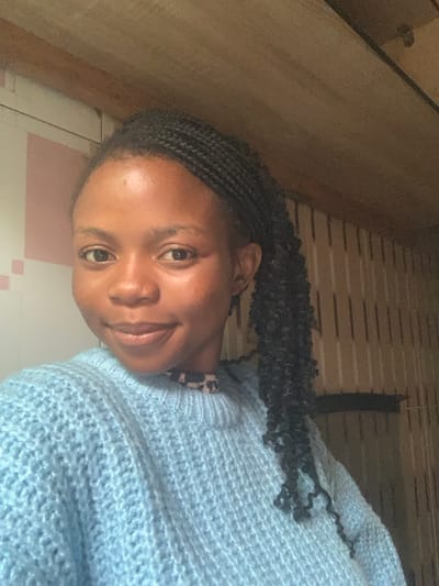 A single individual, an adult female with dark skin, is depicted from the chest up, positioned slightly angled towards the viewer's left. She has dark, braided hair, with some strands cascading over her right shoulder. Her eyes are open and directed towards the camera, and her lips form a slight smile. She is wearing a light blue, chunky-knit, long-sleeved sweater. A black and white patterned garment is visible at the neckline underneath the sweater.
The background consists of two distinct wall sections within an indoor environment. The left portion features a light-colored wall with faint, pale pink rectangular patterns. Above this, a horizontal wooden beam is present. The right portion of the background displays vertical light brown panels, with another horizontal wooden beam running across the top. The lighting is even, suggesting an interior space. The specific location within the city of Onibudo, Nigeria, is not discernible from the visual information provided.