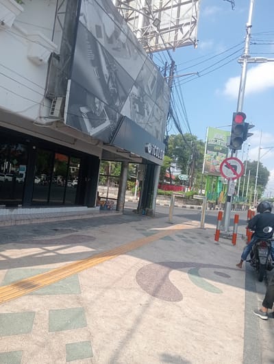 A motorcycle is parked on the side of the road in Sidoarjo, East Java. The motorcycle is facing away from the camera and is partially obscured by a pedestrian. A traffic light is visible in the background, which is red. There is a billboard advertising Indomie noodles in the background as well. The scene appears to be in a commercial area, with several buildings and businesses visible. The sky is blue and clear, with a few clouds.