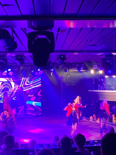 In Hitis, Finland, a dimly lit indoor performance space is illuminated by vibrant purple and blue stage lighting. A large LED screen displays abstract, colorful graphics. On a raised stage, several performers are active. In the foreground, a man in a patterned shirt is on his knees. Further back, three women in red jackets and dark skirts are dancing. The audience is seated in tiered seating in the lower portion of the image, their faces and the tops of their heads visible in the ambient light. The overall scene suggests a lively musical or dance performance.