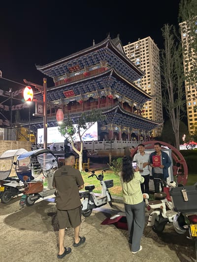 In Eshan Yi Autonomous County, China, at night, a group of people pose for a photograph in front of a traditional Chinese building with ornate blue and red tiered roofs. Two men stand side-by-side, one in a plaid shirt and the other in a striped shirt, holding a certificate. A woman in a light green shirt and gray pants is taking their picture with her phone. The background features tall, modern apartment buildings and trees. Several electric scooters and a covered tricycle are parked in the foreground, casting shadows on the paved ground. A large screen displays an image of a similar traditional building.