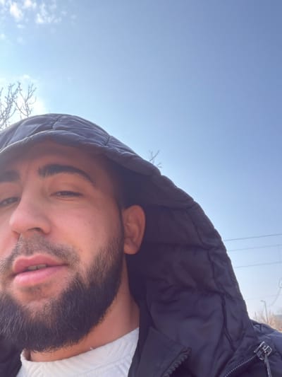 The image features a male individual, appearing to be in his 20s or 30s, occupying the lower half of the frame in a close-up outdoor shot, likely a selfie. He has dark hair, a prominent dark beard, and dark eyebrows. He is wearing a dark, possibly black or navy, puffy hooded jacket, with the hood up, framing his head. A white, ribbed high-neck garment is visible beneath the jacket. His left eye is slightly squinted, and his lips are slightly parted as he looks directly into the camera. The upper half of the image consists primarily of a bright, clear blue sky with minimal, wispy white clouds. In the background, particularly on the left and subtly on the right, bare tree branches without leaves are visible, suggesting a colder season. Faint horizontal utility lines and indistinct distant structures are discernible in the far right background, indicating an outdoor, possibly urban or semi-rural setting. The scene is understood to be in Kermen, Bulgaria, though no specific city landmarks are visually discernible within the image itself.