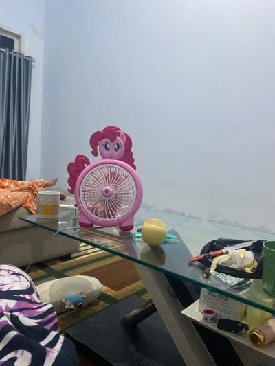 The image shows a room with a glass-top table. On the table, a pink fan shaped like the My Little Pony character Pinkie Pie is visible. Next to the fan are various items, including a green cup, a black plastic bag with crumpled paper and what appears to be a red pen or tool, a roll of medical tape, a small black object that could be a key fob, and a packet of "DISPOSABLE MASKS (NIJAG)". There is also a yellow object that resembles a round fruit. Below the table, a person's legs are visible, clad in an orange patterned fabric. A plush slipper with a small blue object on it rests on a rug. The background shows a white wall and a gray curtain. The lighting suggests it is daytime, possibly late afternoon or evening, due to the ambient light. The overall scene appears to be in a residential setting in Walennae, Indonesia.
