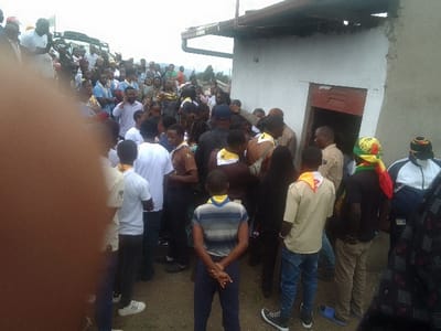 A large crowd gathers near a white building with a red door. Many individuals wear scout uniforms with yellow neckerchiefs. People are dressed in various attire, including white shirts, blue jeans, and patterned fabrics. A vehicle is visible in the background amongst the throng. Some wear head coverings, and the overall atmosphere seems lively, potentially indicating an event or gathering.