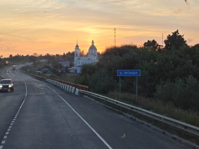 The image depicts a road scene at sunset, with a warm orange and yellow sky dominating the upper portion. A white church with multiple domes and a tall steeple is visible in the mid-ground, nestled among trees and other buildings. A blue road sign with Cyrillic text, "р. МОКША," is positioned on the right side of the road. The road itself is asphalt, with lane markings and a guardrail. A dark-colored car is driving on the left lane of the road, and its headlights are illuminated. The environment appears to be rural or semi-rural, with vegetation and scattered structures. The overall impression is one of a tranquil evening drive.