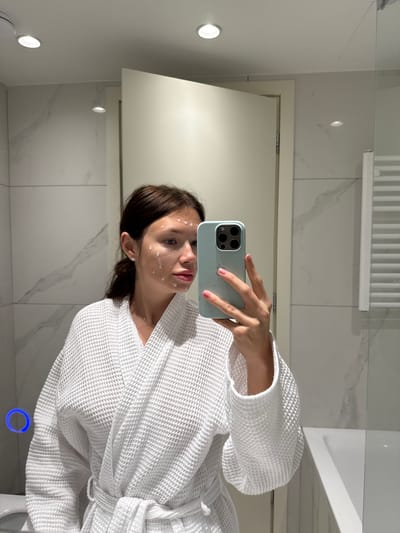 A female subject is captured in a mirror selfie within a modern bathroom located in the Municipal Unit of Elliniko, Greece. She has dark brown hair pulled back and is wearing a white waffle-knit bathrobe tied at the waist. Her face exhibits numerous small, discrete applications of a white cosmetic substance. The subject holds a light blue Apple iPhone, featuring a triple-lens camera array, in her right hand; her fingernails are painted pink. The bathroom environment includes walls covered with large white marble-effect tiles and a white ceiling with two visible recessed spotlights. Reflected in the mirror are a partially open white door with a light-colored frame, a white heated ladder-style towel rail, and the edge of a white bathtub. On the lower left of the mirror, a blue illuminated circular control is visible above a portion of a white toilet bowl.