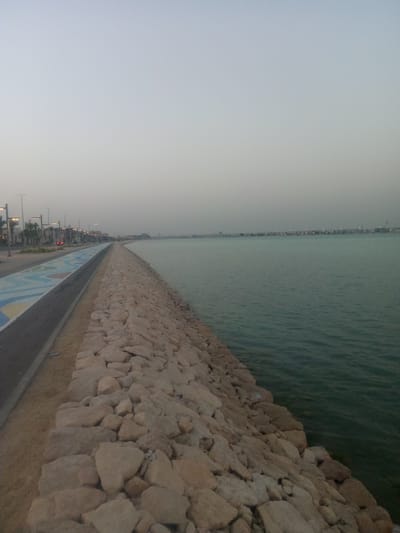 The image depicts a coastal scene in Al Khobar, Saudi Arabia. A wide paved promenade runs along the left side, featuring a decorative painted surface in blue, yellow, and white. Streetlights line the promenade. A rock barrier separates the promenade from the water. The water appears calm and extends to the horizon, where the distant skyline of the city is visible. The sky is overcast.