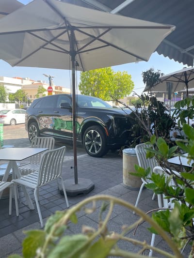 The image displays an outdoor scene in Nes Ziona, Israel, specifically near the intersection of Derech Weizmann and Rothschild Street, indicated by the Winged Victory statue visible in the background. A dark-colored SUV, possibly a Cadillac XT6, is stationary on a paved parking surface. Its front passenger-side bumper and fender area show damage, with a large, snapped tree branch, consisting of dry leaves and twigs, lodged against the vehicle's front. The branch appears to be detached from a larger tree. In the foreground and midground, an outdoor seating area features two open, light-colored patio umbrellas, one centrally prominent. Several white metal tables and matching chairs are arranged on a grey paver patio. A cylindrical, light-toned container, possibly a trash receptacle or planter, is situated near the right-hand chairs. Out-of-focus green foliage from a foreground plant partially obscures the lower portion of the image. The background includes light-colored buildings, other parked vehicles, a "No Entry" street sign, and a large green-leafed tree. The sky is clear and blue, suggesting daylight conditions. No individuals are visible within the frame.