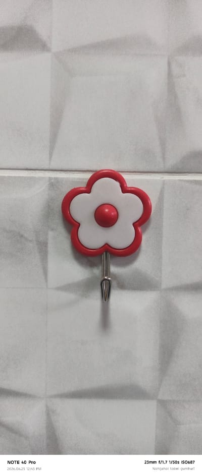 The image shows a close-up, vertical shot of a white and red flower-shaped wall hook against a textured white tiled wall. The hook is centrally placed and made of metal. The flower has five white petals outlined in red, with a red circular center. The background wall has a geometric, slightly textured pattern, with subtle shadows that suggest directional lighting. The image is a still shot, with no discernible activity or event.

Visible text at the bottom left corner reads: "NOTE 40 Pro" and "2026.06.25 12:45 PM". The bottom right corner displays camera settings and some text in a foreign language: "23mm f/1.7 1/50s ISO687" and "Nohijahoi tobei çumhurī". The timestamp "2026.06.25" indicates a future date. The location context provided is Roghun, Tajikistan. The lighting and the plain background suggest an indoor setting, possibly a bathroom or kitchen, given the presence of a wall hook. There are no people visible in the frame, and the overall mood is neutral and objective, focusing on the object and its presentation.