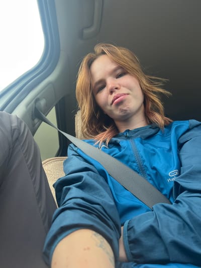 A young woman with shoulder-length auburn hair and fair skin is sitting in the passenger seat of a vehicle. She is wearing a blue zippered jacket and a grey seatbelt. Her expression is pensive, with her lips pursed. To her left, a person wearing dark pants is visible. The background shows the interior of a car, including a window and a part of the dashboard. The image appears to have been taken outdoors, with natural light illuminating the scene. The location is not specified.