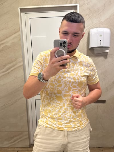 The image shows a young man taking a selfie in a restroom. He is wearing a yellow and white floral print short-sleeved shirt and light-colored shorts. He has short dark hair and a beard. On his left wrist, he wears a silver watch with a green face. He is holding a smartphone in his right hand, which is pointed towards a mirror. The background consists of marble-tiled walls and a white paper towel dispenser mounted on the wall. A white framed mirror is visible, reflecting the man. The overall lighting suggests it is daytime. The location is identified as Gelemenovo, Bulgaria. The man's expression is relaxed, and he appears to be posing for the photo.