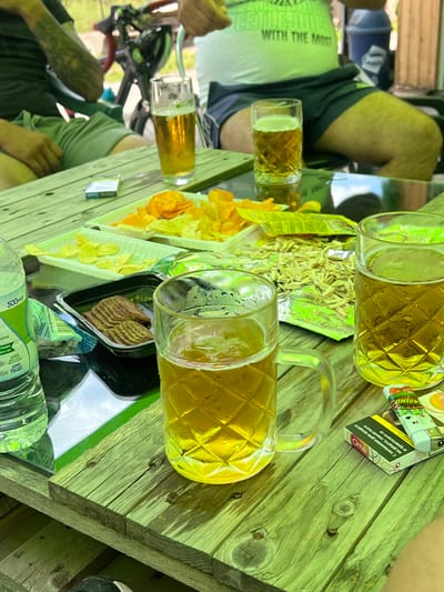 An outdoor scene in Касапите, Bulgaria, depicts a casual gathering around a rustic wooden table under daylight. The table is laden with snacks and beverages.

Three individuals are partially visible. On the left, a person wearing a dark short-sleeved shirt and green shorts displays a prominent tattoo on their left arm. In the upper right, another individual wears a white t-shirt with partially visible green text ("WITH THE MOS") and dark shorts, exposing their legs. A third person's leg and foot, wearing a dark shoe with a white detail, are visible at the bottom right.

The wooden table, stained with a light greenish-brown hue, holds the following items:
*   Three clear glass mugs, each featuring a diamond-patterned texture, filled with an amber liquid, consistent with beer. One mug in the foreground has a handle.
*   A 500ml plastic water bottle with a light green label is positioned on the far left.
*   Two light green plastic trays contain potato chips of varying shades (orange and yellow).
*   A black plastic container holds numerous slices of dark, possibly cured, meat or sausage, arranged in an overlapping pattern.
*   An opened silver and green foil packet, containing spread-out shelled seeds, rests on the table.
*   A white cigarette pack, marked with "Omé" in red and Cyrillic script, is paired with a white lighter adorned with green and red "OASINO" text. A second blue and white cigarette pack is visible further back on the table's left side.

The background shows a bicycle with red handlebars on the left and a blue cylindrical trash can on the right, suggesting an outdoor recreational or picnic area.