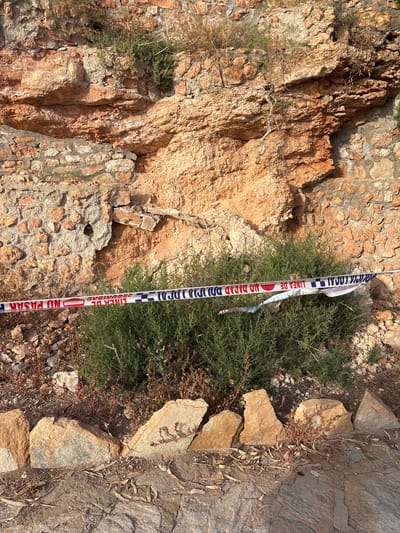 The image depicts a section of a rustic, stone and brick wall with weathered textures, likely part of an outdoor structure in Urbanización Cabo Roig, Spain. A police or security cordon tape, featuring red, white, and blue stripes with Spanish text that appears to read "LINEA DE SEGURIDAD" and "NO PASAR," is stretched horizontally across the foreground, obscuring a dense patch of green shrubs and dry vegetation. The tape is partially torn and appears to have been secured in places with additional tape. The ground in front of the wall is covered with loose stones, dirt, and fallen leaves, with larger, weathered rocks forming a boundary in the lower portion of the frame. The overall scene suggests a restricted or cordoned-off area, possibly due to an incident or ongoing investigation.