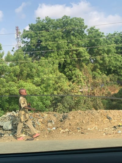 The image depicts an outdoor scene in Abobo, Nigeria, under a bright, partly cloudy sky. A young boy, appearing pre-teen with closely shaved hair, walks from left to right across the frame. He wears a patterned beige and brown short-sleeved top and matching long trousers, paired with pink slip-on shoes. In his left hand, he holds a dark, cylindrical object. The boy is positioned alongside a paved or dark surface, observed from what appears to be a vehicle's window frame in the foreground. Adjacent to his path is a strip of light brown earth, characterized by several small mounds and extensive litter, including plastic bags, paper scraps, and other refuse. Behind this debris and extending into the background is dense, lush green foliage, composed of numerous trees and bushes. A multi-level metal lattice telecommunications tower rises above the tree line on the left side of the image. Several horizontal utility lines are visible across the upper portion of the scene, partially overlapping the trees and sky.