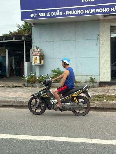 A man wearing a blue helmet and a blue sleeveless shirt rides a black and yellow motorcycle down a paved street. He has on black shorts with red stripes and is wearing sandals. The motorcycle has a white and yellow sticker on its side. In the background, a building has a sign with Vietnamese text, which appears to be an address: "ĐC: 508 LÊ DUẨN - PHƯỜNG NAM ĐÔNG HÀ." The building also has a small shrine with a red and gold roof on a pillar. There are some white flowers planted in front of the shrine. The street has a white painted line. The sky is overcast.