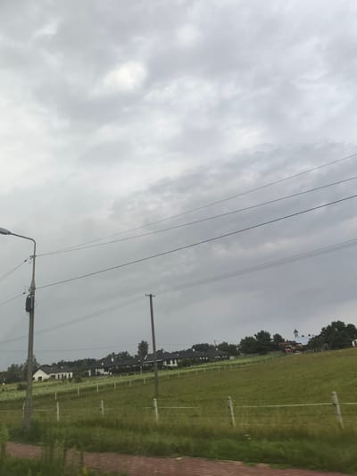 The image captures a cloudy sky over a rural landscape near Skrzeszew, Poland. In the foreground, a brick-patterned path runs horizontally, bordered by green grass and a low wire fence with wooden posts. A lamppost stands to the left, and further to the right, another utility pole is visible, both with electrical wires extending across the frame. Beyond the fence, a gently sloping green field is dotted with more fence posts. In the middle ground, a row of houses with dark roofs is nestled among trees. The sky is overcast with shades of gray, with some lighter patches suggesting diffused sunlight. There are no people visible in the image.
