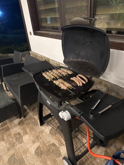 The image shows a black gas grill with its lid open, located on a tiled patio. Skewers of meat and sausages are cooking on the grill. Dark wicker chairs surround the grill. Through a window in the background, a dark evening scene is visible, which possibly indicates that this image was taken in the city of Pchelin, Bulgaria, as requested. There are no people present in the image.