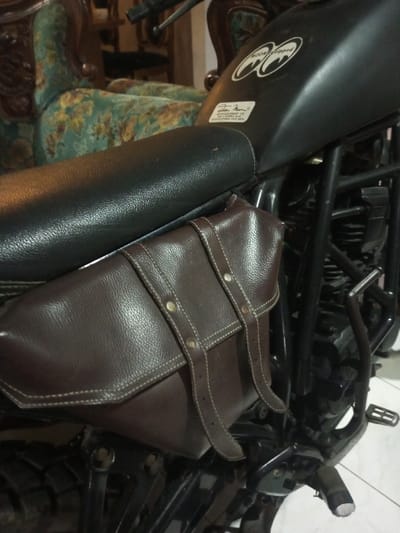 A black motorcycle is depicted indoors on a light-colored tiled floor in B. Nam Ngao, Laos. A dark brown, textured faux leather saddlebag is securely attached to the left side of the motorcycle, below the seat. This saddlebag features two vertical straps with light-colored stitching and metallic fasteners. The motorcycle has a black textured seat and a black fuel tank, which displays a white "MOON EQUIPPED" logo sticker and a rectangular white label with black text beneath it. Visible motorcycle components include sections of the black frame, engine, a footpeg, and a portion of a knobby tire at the bottom left. The background is blurred, showing ornate wooden furniture, specifically an armchair with green and floral upholstery, indicating an indoor environment. No individuals are present.