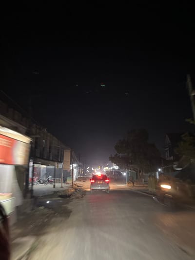 This media file captures a nighttime scene on a street in Landuh, Indonesia, likely taken from a moving vehicle. The image is oriented vertically, with the road and the right side of the scene appearing blurred due to motion.
The scene is characterized by artificial lighting from streetlights and buildings, illuminating a wet road surface that reflects the lights. On the right side of the road, there are buildings, some with signs, and parked motorcycles. A silver car with its brake lights illuminated is visible ahead, suggesting traffic. Trees are also present along the side of the road. The left side of the image is predominantly dark, with very faint lights visible in the distance and some blurry green and red lights in the lower left corner, possibly traffic signals.
The time of day is clearly night, and the wet road suggests it might have recently rained or is still raining, indicating damp weather conditions. The presence of buildings, motorcycles, and cars points to an urban or semi-urban setting in Indonesia. There is no discernible text in the image that can be clearly read. The overall impression is a typical nighttime street view in an Indonesian town, with the motion blur adding a sense of movement and dynamism to the scene.